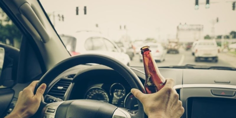 Hands on car steering wheels with one hand holding a beer